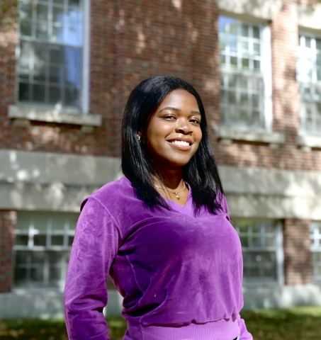 Arianna smiling in a purple sweater and posed in front of a brick building.