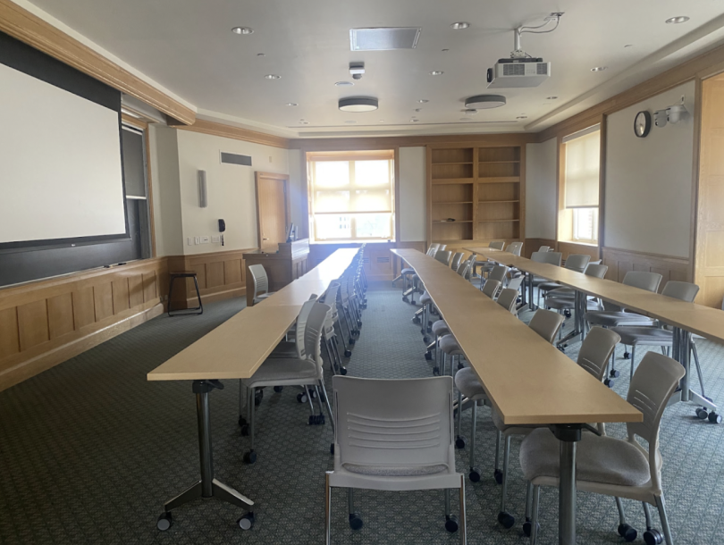 A traditional classroom with long wooden tables, gray rolling chairs, a projector screen, and large windows letting in natural light.