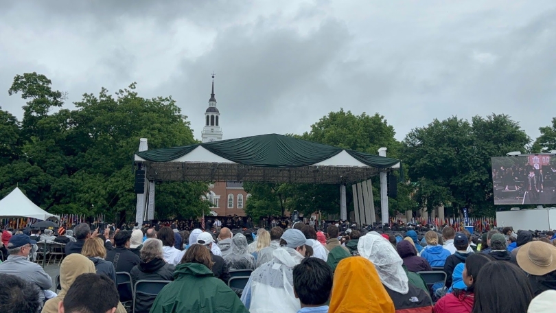 A large stage on the Green with Baker Tower in the background. In the foreground, there are many rows of people with raincoats on. The sky is cloudy and gray.