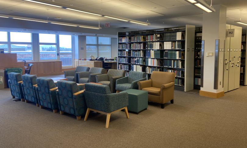 Five blue cushioned chairs sit opposite five brown. Behind the chairs, there are bookshelves filled with books. On the left, there are windows with sunlight shining through.