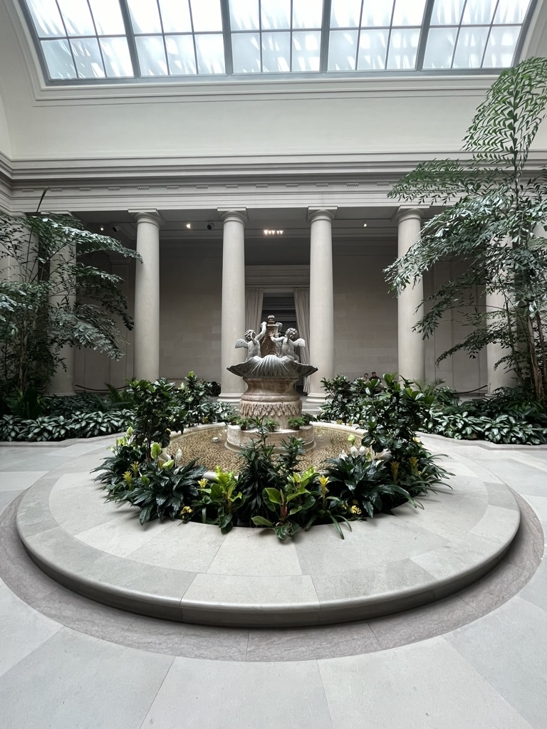 An indoor grotto featuring an archaic fountain. Trees and natural light frame the picture, with the fountain in the center. Big columns are in the back, making the structure look very formal.