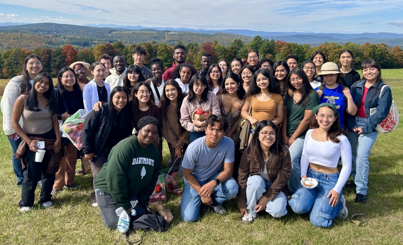 A large group of smiling first-generation college students poses together in front of a scenic landscape with vibrant autumn trees, holding bags of freshly picked apples.