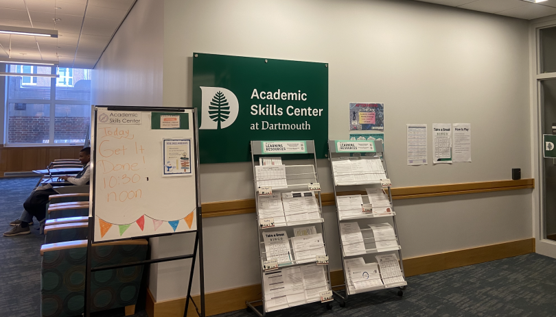 A wall with a green sign that says "Academic Skills Center at Dartmouth." There is a shelf with a white paper pamphlet and a whiteboard with illegible writing. 