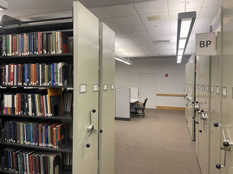 A grey bookshelf takes over the left half of the screen. In the background, you can see a wooden chair and a desk. The floor is brown carpet, and the ceiling is white.