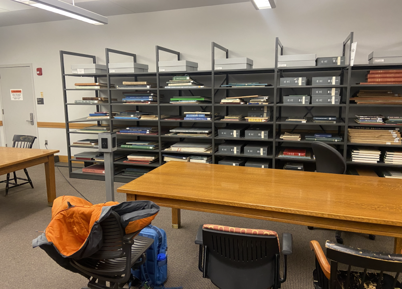 A brown wooden table is in the middle of the photo. Behind it, there are sleeves with oversized books, and next to the table, there is a chair with an orange jacket.