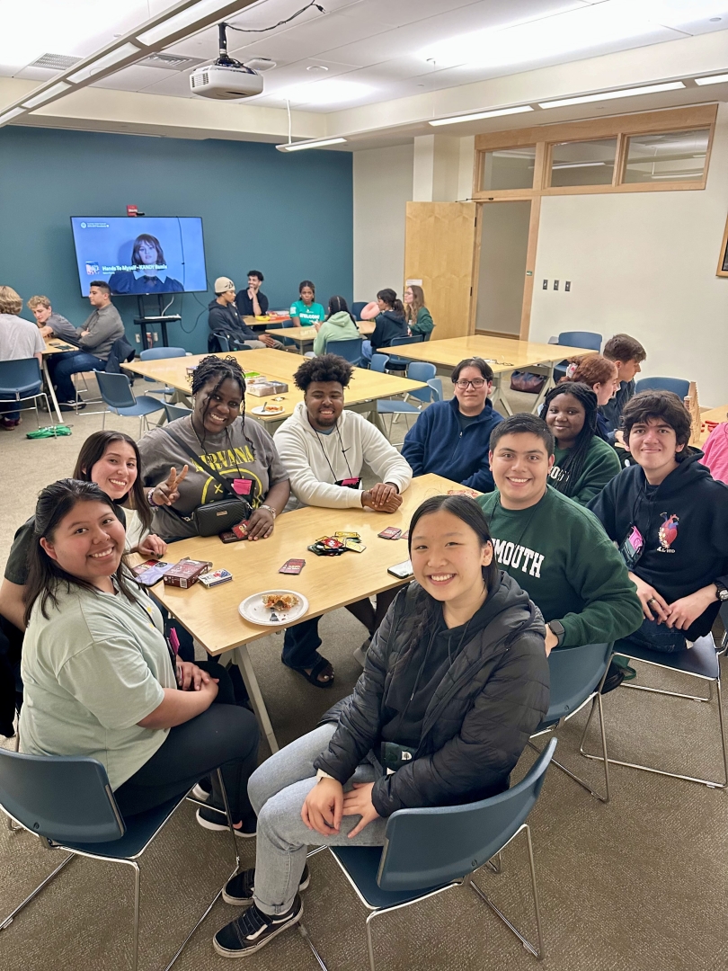 A group of admitted students sit around a table playing card games and eating pizza indoors.