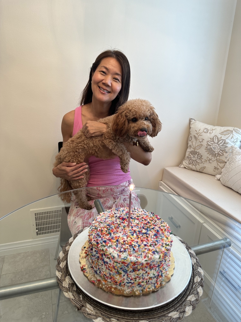 My mom holds my small brown dog Kelsey and smiles at the kitchen table with a rainbow sprinkled birthday cake.