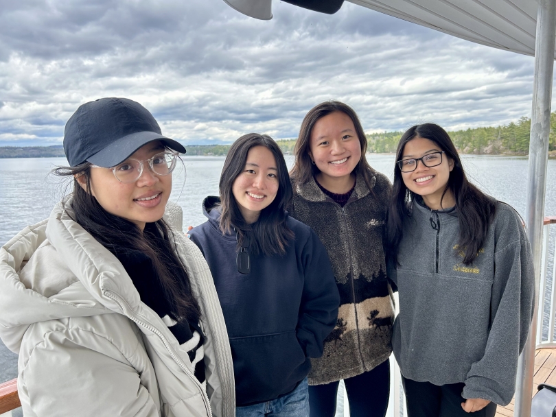 Four girls smile at the camera in a horizontal shot with Lake Sunapee in the background on a cloudy day.