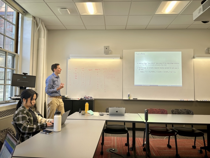 An economics professor with a blue shirt lectures to the class about fixed effects using a slideshow and the whiteboard.