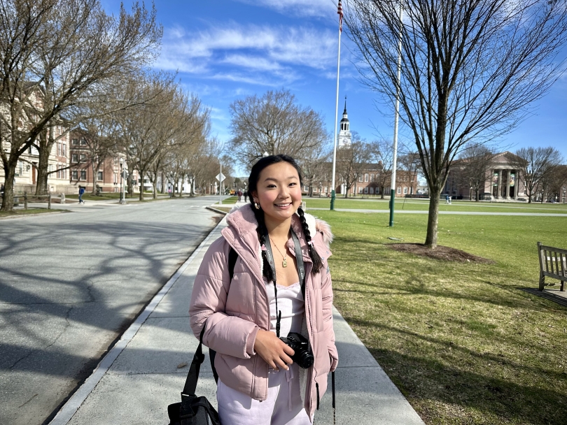 My sister is wearing a pink coat and standing on the sidewalk near the Green with her camera around her neck.
