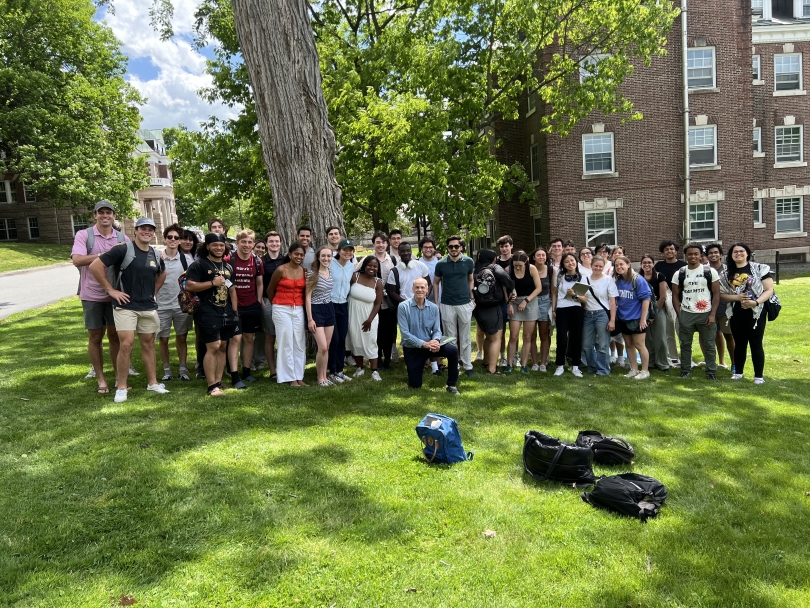 A group of physics students smiles for a picture around a professor on the grass near a tree.