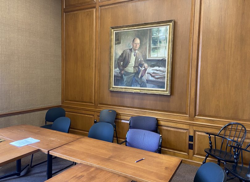 A wooden table with blue chairs sits in the forefront of the image. On the back wall is a painted portrait of a man sitting on a desk and holding a book.