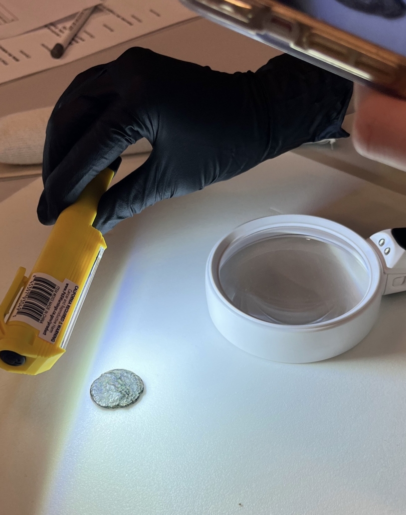 An ancient coin on a white table. A gloved hand is holding a light above the coin, and a magnifying glass is sitting on the table next to the coin. 