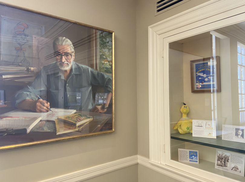 A framed portrait of an old man hangs on the left wall. The man has gray/white hair, is wearing a blue shirt, and is writing in a boom. On the right, there is a display case with a yellow plush and some printed cards.