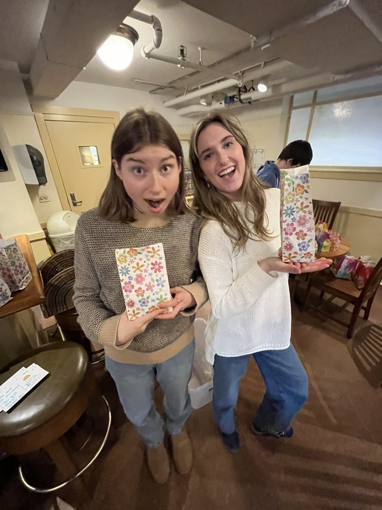 Two young women smile in sweaters. They are both holding goodie bags in their hands.