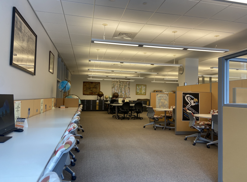 A large room with desks and laptops lining the left side. On the right, there are small cubbies. On the back walls, there are paper maps and globes.