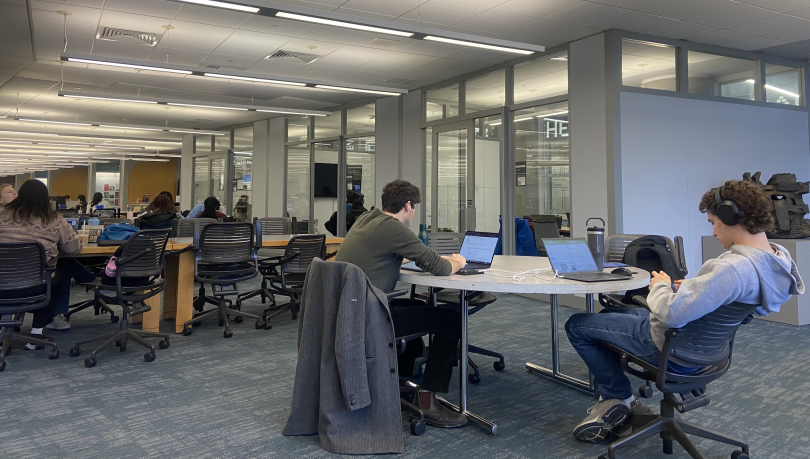 Students are studying at a desk, and in the background, there are grey study rooms. The floor is carpeted, and the tables are gray