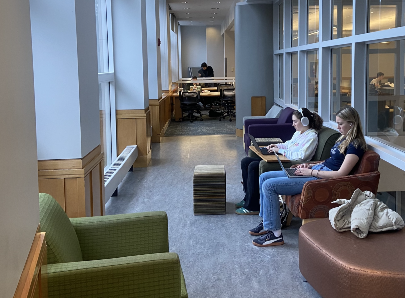 A carpeted hallway with windows on both sides. Cushioned chairs on either side. Two students sit on the right side of the hallway and are using their laptops.