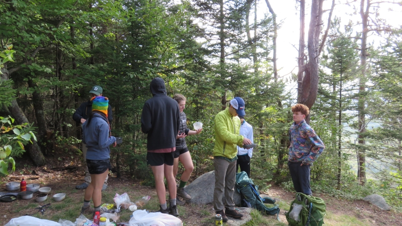 An image of me, Crystal, and my trip group outside at a campsite. We are all standing and in the middle of packing up our camping tools. The background is a forested area and there are large rocks in the center of our campsite.