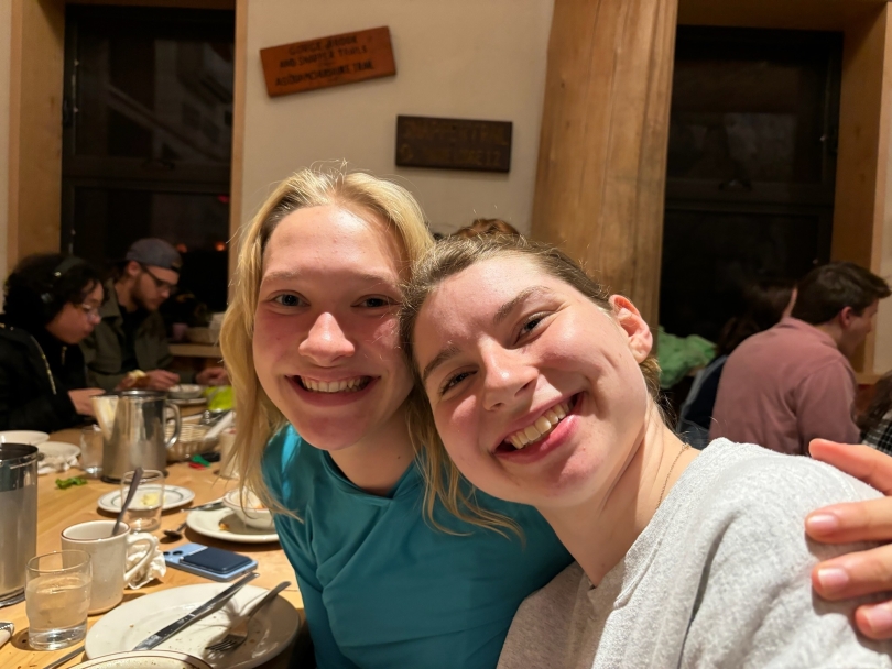 Two white girls smile for a photo while sitting down for dinner at the Moosilauke Ravine Lodge. One is wearing a blue sun hoodie and the other is wearing a grey sweatshirt.