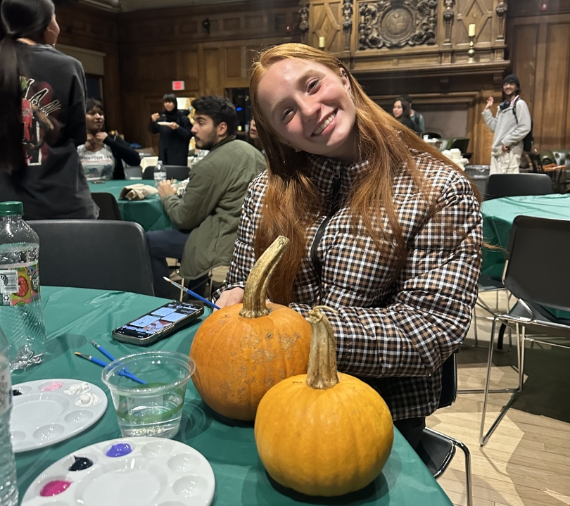 A first-generation student smiles while posing with an unpainted pumpkin at a community dinner. The table is covered with painting supplies, like palettes.