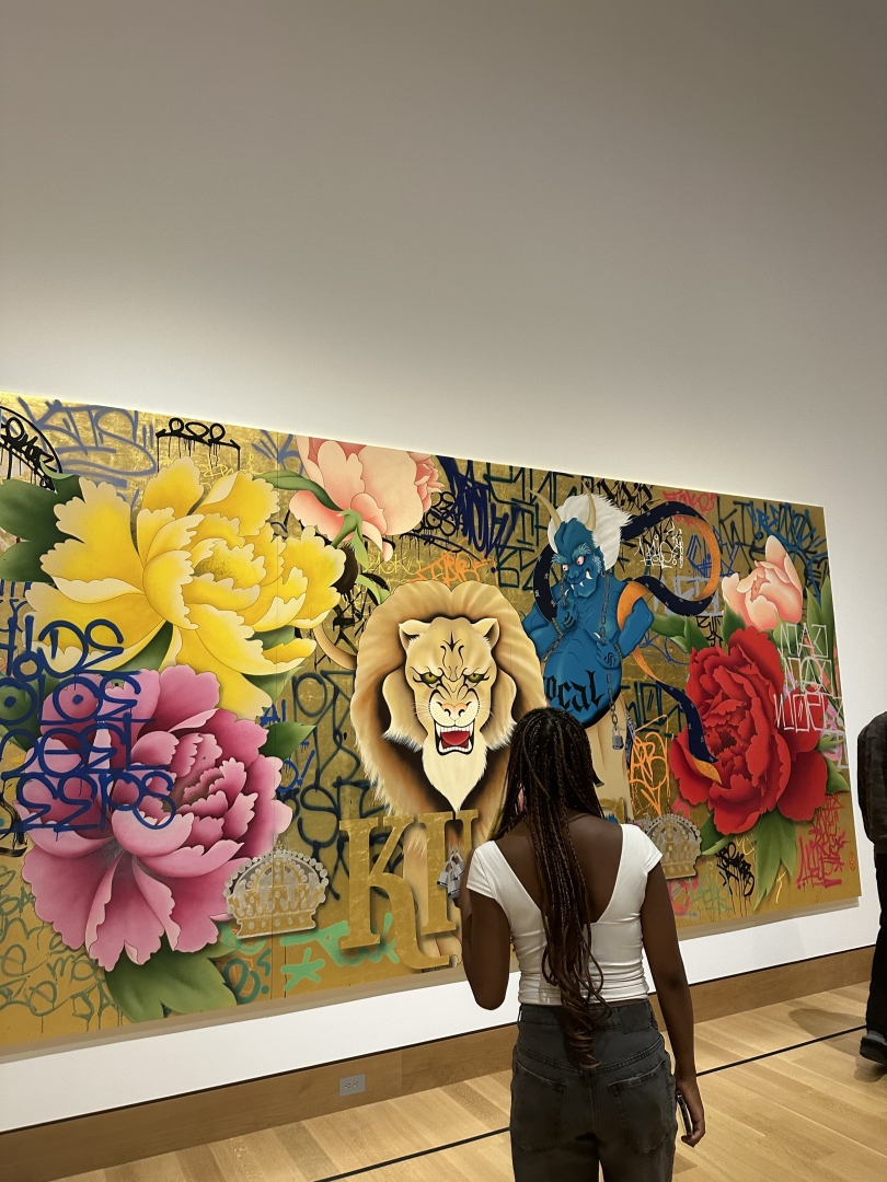 A girl with curly braids stands in front of a colorful portrait on a blank, white wall.