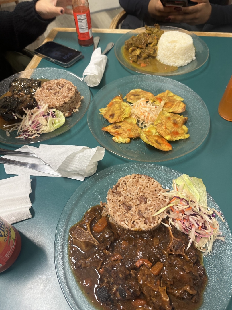 A plate of Jamaican food featuring oxtail, rice and peas, plantains, and coleslaw, served on clear plates on a blue table.