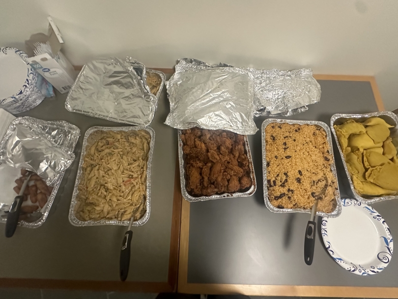 Five large aluminum trays filled with Jamaican cuisine sit on a table, showcasing golden fried festival, creamy rasta pasta, crispy chicken wings, rice, and flaky chicken patties.