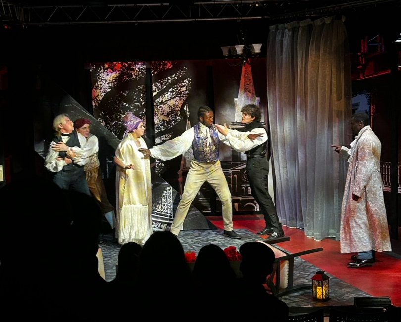 A dynamic scene from BUTA's rendition of Hamlet features a few actors in dramatic poses, their expressions brimming with intensity. The dimly lit stage is accented by minimalist props and evocative lighting, drawing focus to the performers.