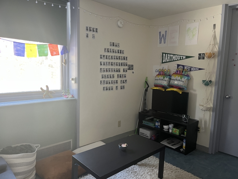A room with white walls and a window. A coffee table, white carpet, and TV on a stand furnish the room. The walls are covered in polaroids and posters.