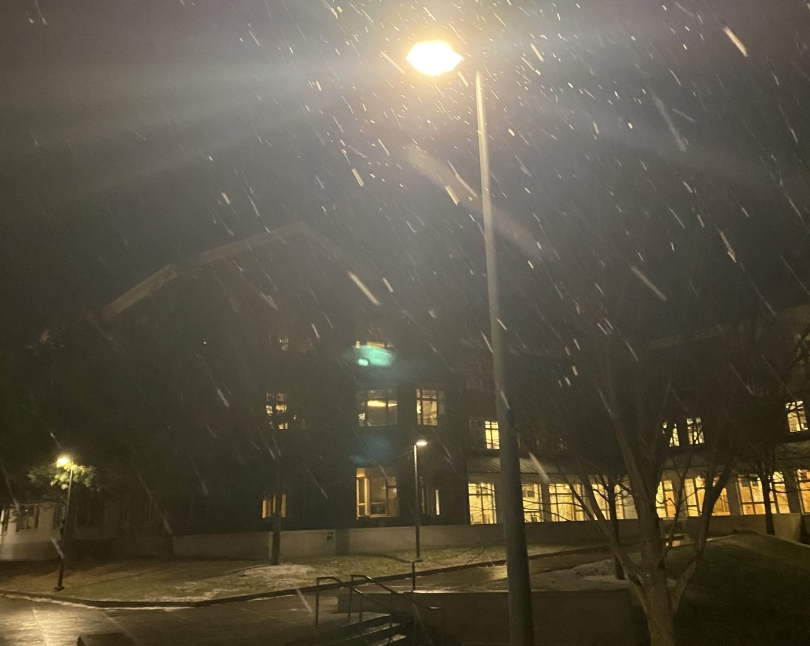 a picture of a lamp post, as clumps of white snow fall around it in all directions, with carson hall in the background