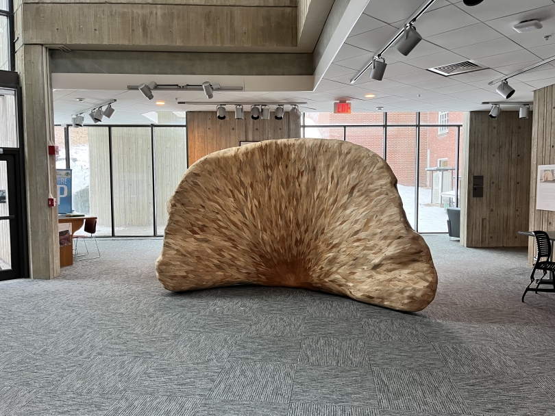 A large, wooden sculpture in the shape of a semicircle. It is composed of thousands of slivers of wood in different colors