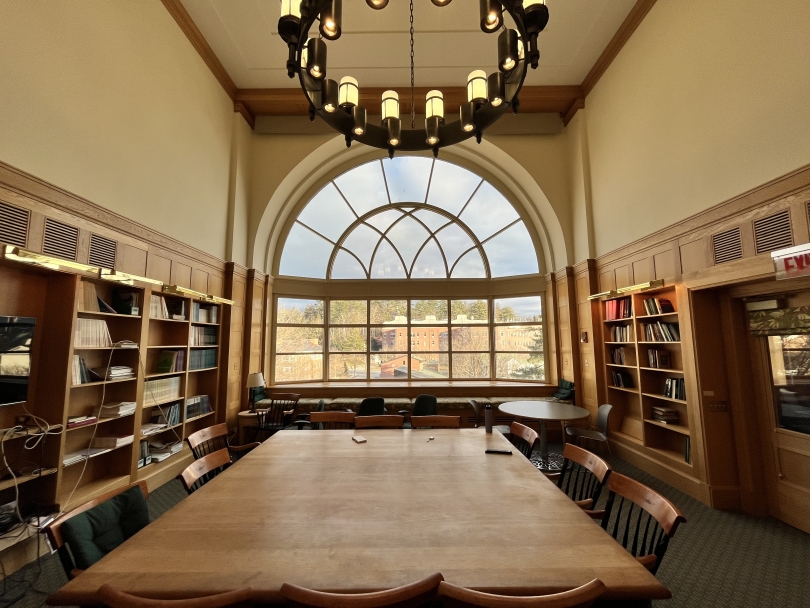 A view out of a massive semi-circle window. In the surrounding room, there is a large wooden table surrounded by chairs, and a variety of wooden bookshelves