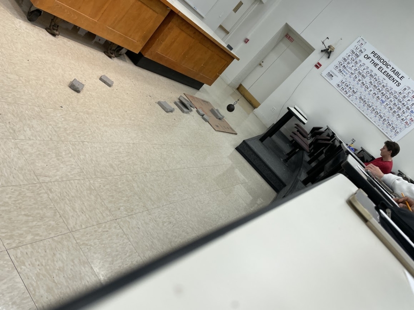 a funny-angle picture of a the broken pieces of a brick lying on the floor under a heavy cannon ball suspended by a cord from the ceiling in Kalina's physics classroom