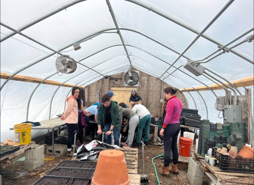 A group of people on the inside of a small plastic greenhouse, using a metal tool to make blocks of soil.