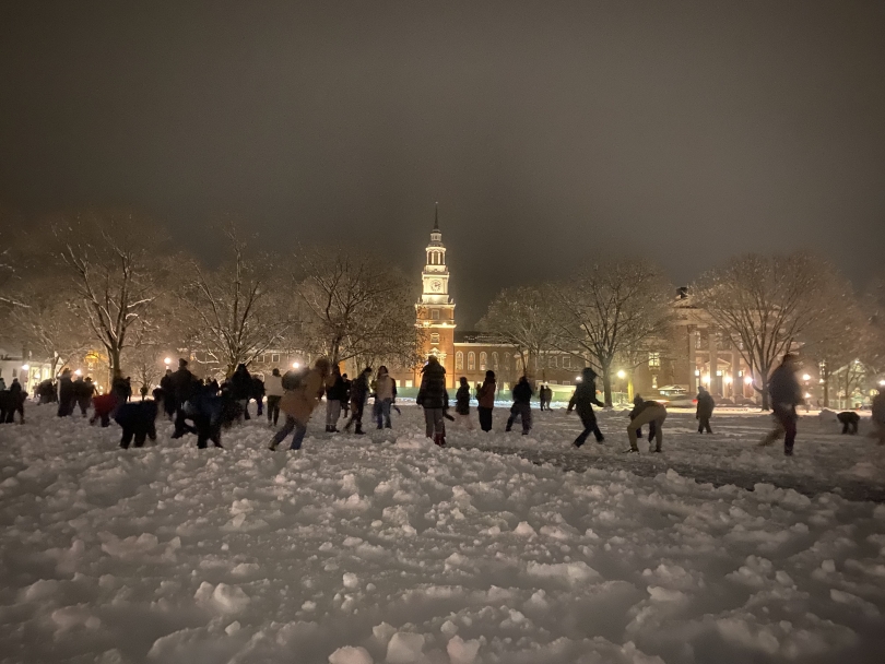 a picture of the snow-covered green at dartmouth with people on it throwing snowballs at each other, with baker-berry library in the background!
