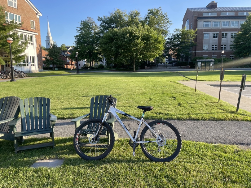 A picture of a sunny evening on Dartmouth's campus near Goldstein Hall with a silver mountain bike propped up against a green Adirondack lawn chair in the imminent sunset