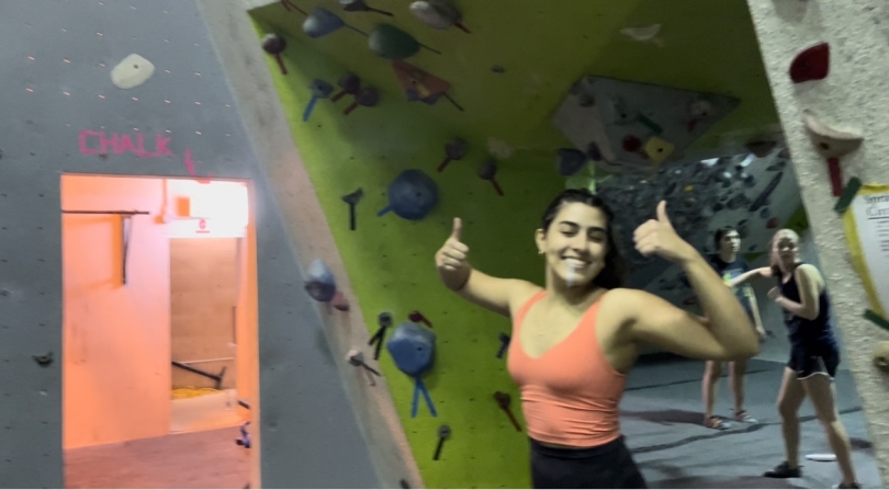 Girl in orange shirt giving a thumbs up by the climbing wall.