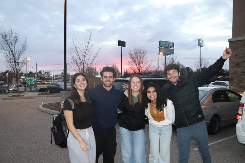 five people standing in a hotel parking lot posing for a picture. The sun is setting in the background
