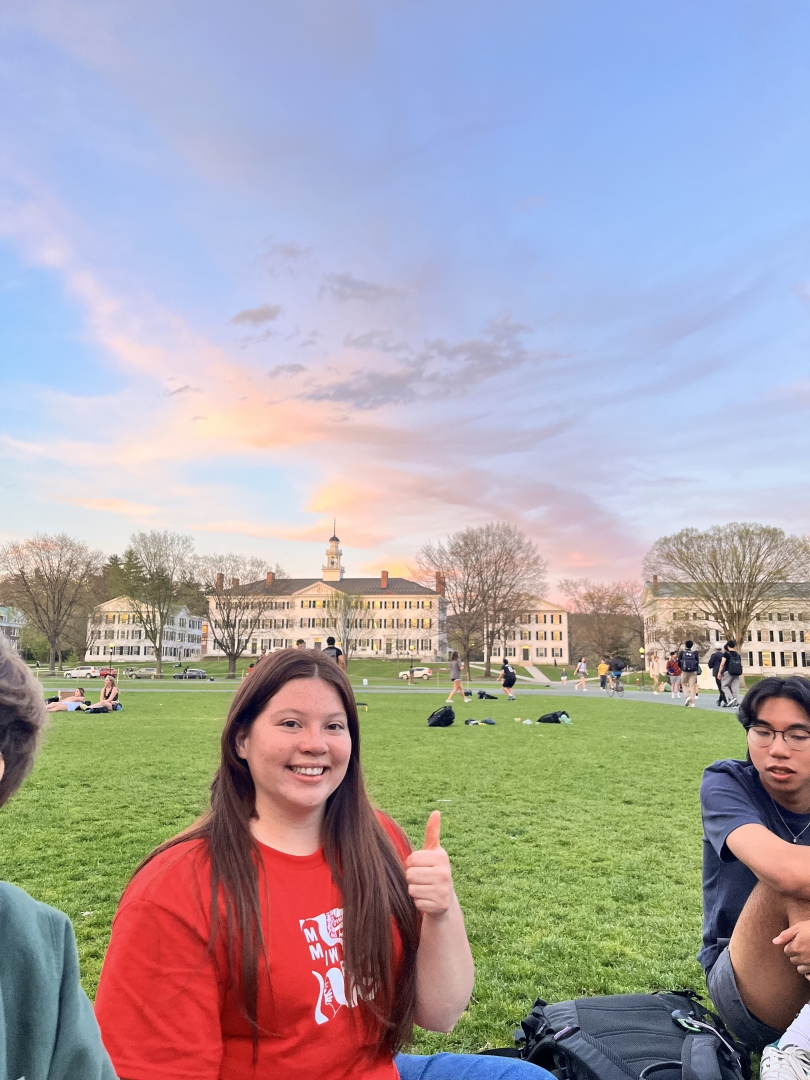 A photo of my friend, Carolina, smiling on the Green. There is a sunset above Dartmouth Hall in the background.