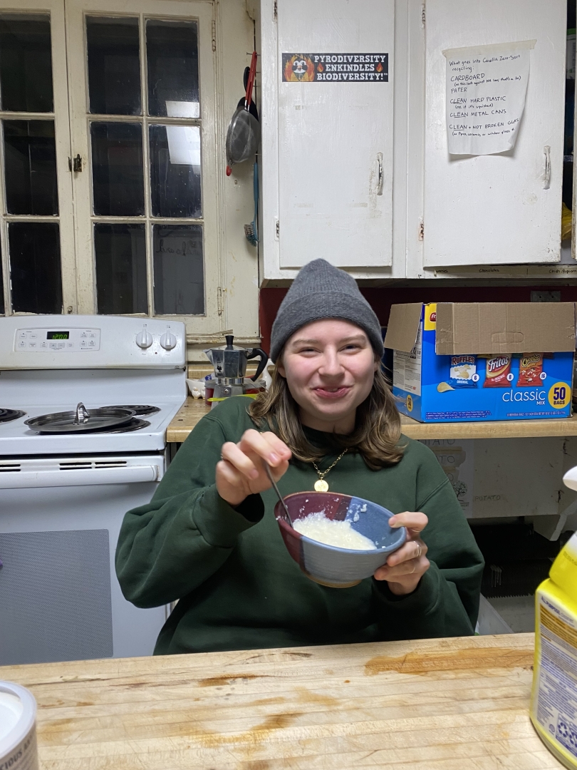 A girl laughs in a kitchen with a bowl of grits. She is wearing a green sweatshirt and a grey beanie.