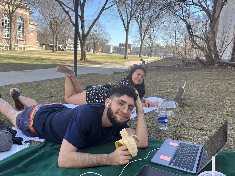 An image of two students laying down on the grass outside Baker Library and studying in the sunshine. 