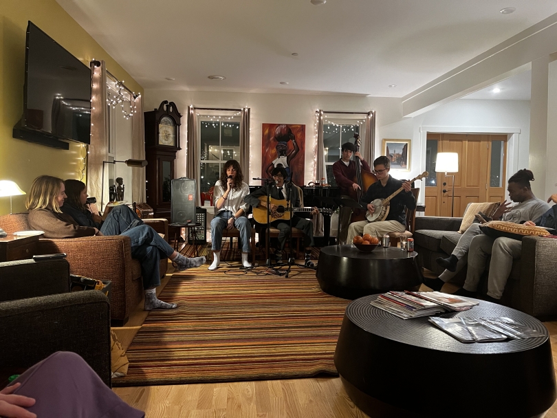 A photo of a bluegrass band performing in a warmly-lit home with an audience