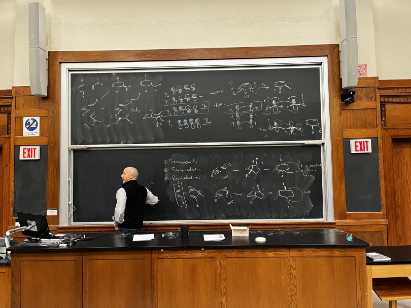 An image of a chalkboard with a man standing in front of it drawing organic molecules on the board.
