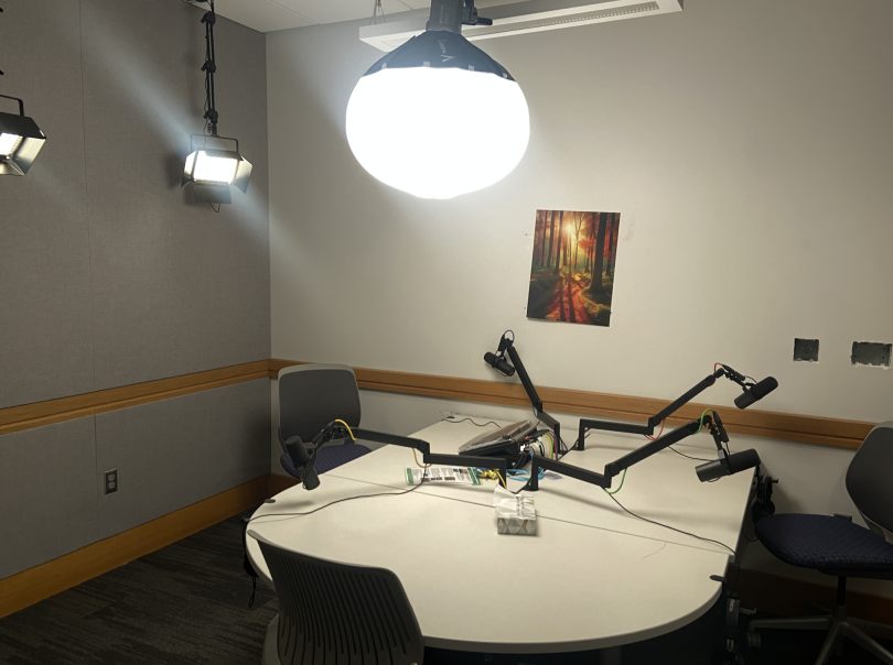 A dark room with a white circle table in the center. There are four chairs and four podcast microphones.