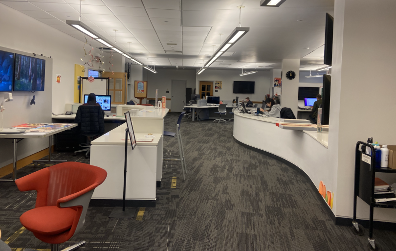 A dark carpet room with a long desk on the left and small tables on the right. There is an orange desk chair in the foreground and a TV mounted on the left wall.