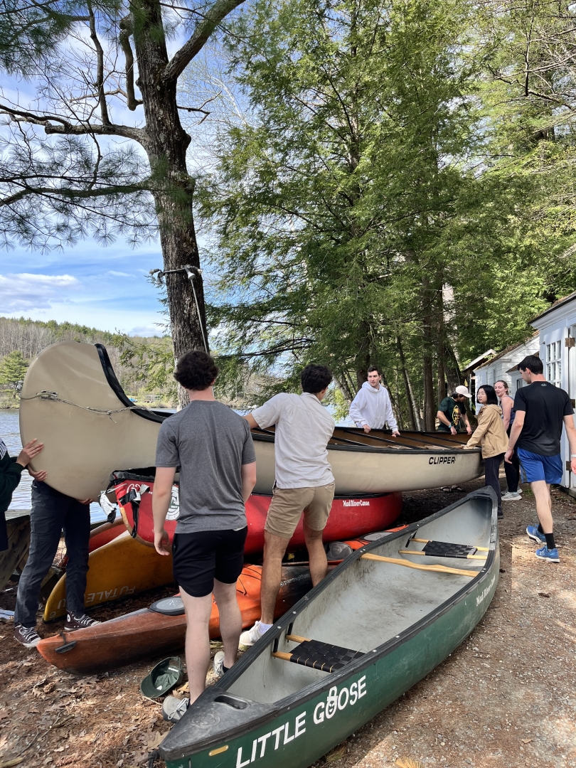 Several students work together to lift a large canoe. There are other canoes and kayaks resting on the ground near their feet.