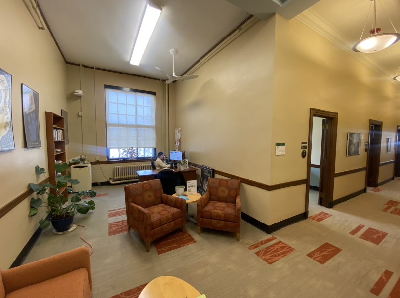 A reception desk sits on the back left, and a hallway lies on the right. The hallways lead to many offices with open doors. There are orange couch chairs in the forefront, and the carpet is a brown and orange pattern.