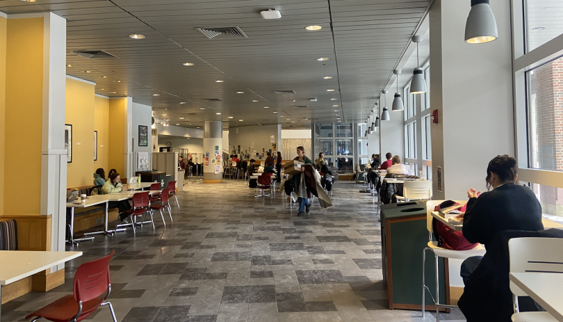 Large open space with yellow wall on the left and windows on the right. There are white tables with red chairs on the edges.