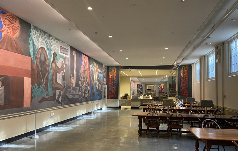 A long hall with overhead lighting and gray tile flooring. On the right, there are long wooden tables with lamps for studying. On the left, the walls are painted with orange, red, and blue paint to create enthralling images of people and architecture.